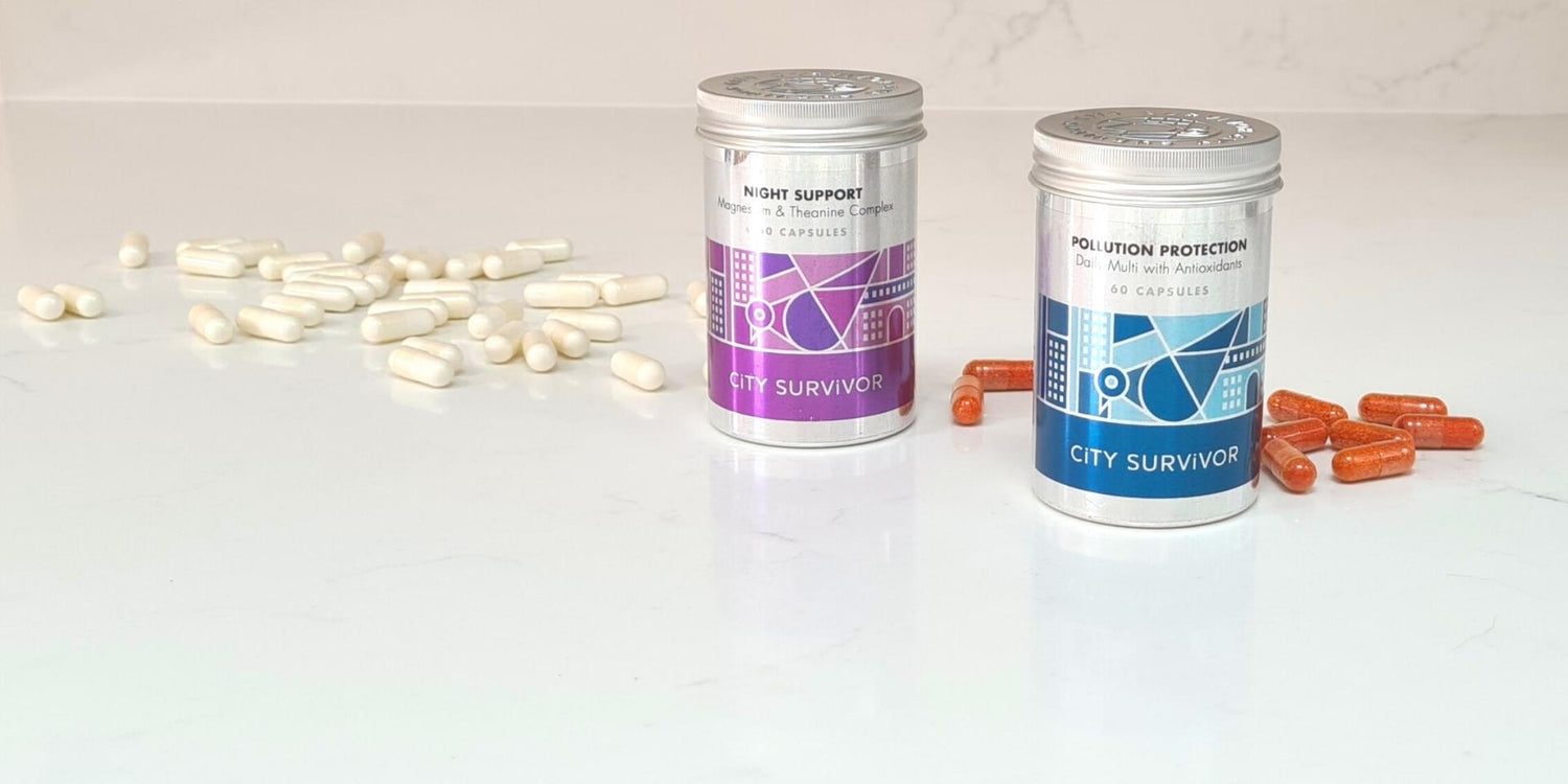 Two metal supplement pots stand on a white marble surface with orange and white capsules scattered around. The product labels read City Survivor Night Support and City Survivor Pollution Protection